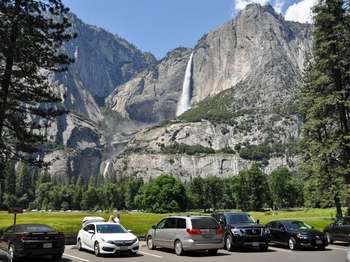 A line of cars are parked in a parking lot next to an open meadow that showcases a view of a tall waterfall.