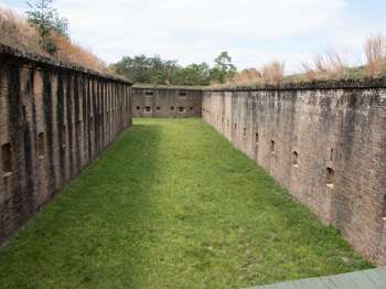 Exterior brick walls of the fort showing loopholes spaced evenly along each wall. A large, open, grassy field is in between the walls.