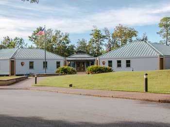 A blue building with a flag pole and grassy hill in the foreground and green trees and blue sky in the background.