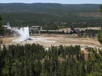 View from the side of a hill showing an erupting Old Faithful Geyser, people watching, and lodges in the background.