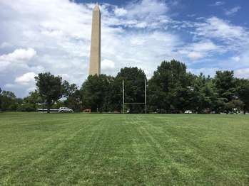 A rugby field surrounded by trees with the Washington Monument towering in the background.