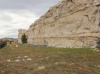 A small cemetery lies next to a tan and grey, 90 degree, highly cracked, solid rock, edge of a cliff