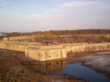 Dark water flooding around the brick structures of the fort in the foreground. A black cannon is mounted on top of the fort. Green trees and blue sky in the background.