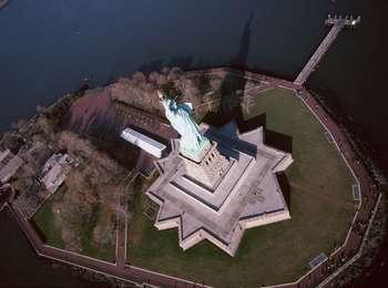 An aerial view of Lady Liberty sets her star shaped base in stark relief against the green grass and blue water surrounding the statue.