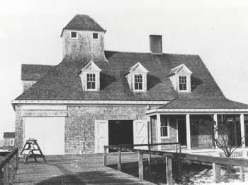 A black and white photo of a white wood sided two-story building with many windows and white doors. A wooden boardwalk in the foreground. Two people standing on the left in the background.