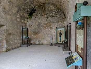 The American Indian Room can be accessed from the courtyard, the Chapel or Room 16. This audio description is from the courtyard entrance. Double wooden doors open into the room. Above and flanking the door are windows with iron bars and wooden shutters that also open inwards. The room is about 20 feet wide and 30 feet long, with an arched ceiling about 17feet high. An entrance to the Chapel is on the west wall. The entrance to Room 16 is on the east wall. Moving from left to right from the entrance, the exhibits, in English and Spanish, include: Osceola. 1804 – 1838. The text explains that Osceola resisted the United States’ policy of forced American Indian removal. He urged Florida Seminoles to fight for their lands. Over his black hair, he wears a burgundy turban, with a large gray feather at the back. He has an angular face. He has a black piece of cloth loosely tied around his neck. Over a long blue patterned shirt, he wears a wide pink scarf with a flower print. He wears an open blue jacket with silver cuffs and ribbons tied at the wrists. Around his waist is a multicolored woven belt. He wears several beaded necklaces, and one with three large silver crescent shaped medallions. He has bag strapped across his chest. His legs are covered with red socks with wide beaded bands, tied at the calves. He holds the barrel of a musket in his hand. Kiowa Sun Dance Exhibit. A vertical glass panel protects a diagram of circles with arrows, carved into the wall by the Kiowa Indians. Text describes symbolism connected to the Kiowa sun dance. Captain Richard Henry Pratt. 1875 – 1878. Text explains that Captain Pratt was assigned to guard the 74 American Indians prisoners at the Castillo and created a program of assimilation for them. An illustration shows him with short brown hair with a large, straight nose, pointy mustache, and goatee (a beard only on the tip of his chin). He wears a blue uniform with gold trim, epaulets, and belt. His pants have a yellow stripe down the outer side. In one hand, he holds an ornate helmet with gold trim and cascade of golden fluffy material at the top. The other hand rests at his side, on the hilt of his sword. A Coquina Canvas Exhibit. A vertical glass panel protects an outline of an Apache Fire Spirit carved into the wall. Text and photos explain symbolism. United States Takes Over Exhibit. An illustration shows Spanish and American soldiers. A photo shows captured American Indians. The text explains that in the 1800s, Indian wars in both Florida and the west saw the U.S. government locking up American Indian prisoners here.