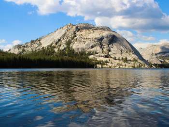 A granite dome reflects into a large body of water. Large puffy clouds dot the sky.