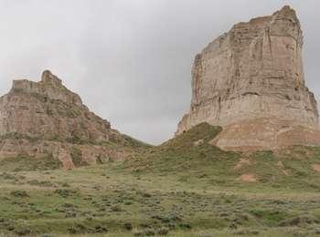 Landscape view of two large, solid rock buttes with sagebrush and grass in the foreground