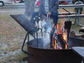 Black fire ring with flames coming out of the top. A man holding a dog near his knees sits on a picnic table in the background.