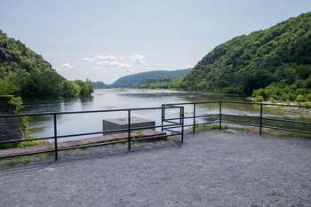 This is a scenic image of the meeting point of the Potomac and Shenandoah. A massive river runs through mountain ranges on either side of it. The mountains are lush and green and stretch off into the distance. A metal bar crosses the front of the river to protect people from falling in.