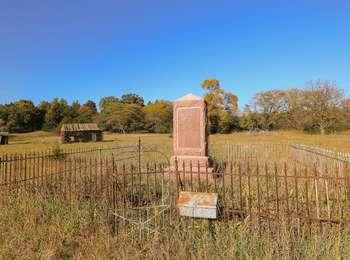 A rusted, metal picket fence surrounds a rock monument in a grass meadow bordered by trees
