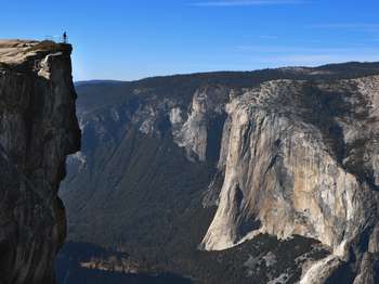 A person stands behind a railing at a cliff's edge, across a valley from a large rock formation that is El Capitan. 
