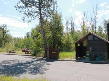 Cottonwood Creek Picnic Area overview with vault toilet, trash and recycling, four picnic tables with visitors sitting at one of them. Cottonwood trees surround the area