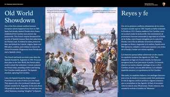 On the far left and right sides of the panel, white text in English and Spanish reads:  Old World Showdown. One of the first colonial conflicts between European nations happened near here in 1565. Spain had already claimed Florida when France established Fort Caroline near present-day Jacksonville. This French outpost threatened the security of Spanish treasure fleets that sailed along the Florida coast. To defend its claim, protect its fleet, and defend the Catholic religion, Spain sent 800 sailors, soldiers, and civilians to remove the French Protestants (Huguenots) from Florida and start a Spanish colony.  The French had barely survived a year when the Spanish founded St. Augustine in 1565. To secure their place in the New World, the French sailed south to attack the settlement. A storm scattered the French ships, stranding soldiers and leaving Fort Caroline weakly guarded. The Spanish attacked, capturing Fort Caroline.  Later, the Spanish found the shipwrecked Frenchmen just south of where you now stand. They spared only a few professed Catholics and artisans needed in St. Augustine. In the end, about 250 people lay dead. Since then, the inlet has been called Matanzas, meaning, â€œslaughterâ€, in Spanish.  The Spanish found the Frenchmen helpless and unable to cross the inlet.  End of Text  In the center of the panel, a color illustration. On a white sandy dune overlooking blue water, a man in a red uniform with gold stripes looks down on a group of men in loose white shirts and brown pants; many of them barefoot. Their heads hang down and their hands are tied behind their back. Behind the man in red, other men, their hands also tied, are pushed forward by soldiers wearing breastplates and silver helmets. On a dune behind these men stands a man in a long black robe with a cross around his neck; his face turned up toward the sky, his hands folded across his chest. To his left, are more soldiers. One in a black, thigh-length jacket and sword looks down on the shackled men. Behind him stand more soldiers looking out over the water; one holds a large red flag.   The panel is set in the elbow of the wooden walkway. Directly beyond the panel is a large oak tree, its branches stretch outward to the left and right. Beyond the tree, on the ground below, thick palm fronds and other vegetation.    [End of message]