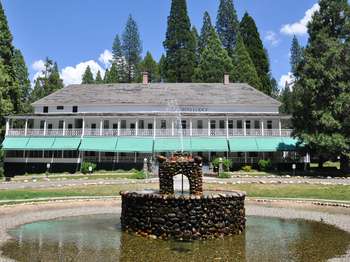 A two-tiered water fountain surrounded by a circular pond is situated in front of a white, two-story building with a dual level, wrap-around porch. 