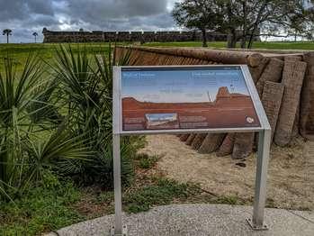 On the top half of the panel is a color illustration of the Cubo Line, an earth and palm-log embankment used to defend the city of St. Augustine. At the left, elevated land leads down to cleared land and a 5-foot deep moat. To the right, just before the rising embankment, a vertical brown stick marks the palisade wall of pine stakes that stood in the moat. To the right, the 9-foot high embankment, made of reddish-brown dirt and palm logs hid soldiers from the invaders’ view. Below, extending from the left side to the pine stake, a white, horizontal line and text in white: “Outside the Cubo line: countryside.â€, To the right, just past the pine stake, a second white horizontal line and the text: “Inside: the protected city.â€, Above the reddish-brown land, a pale blue sky. Along the top of the panel, at left and right, black text in English and Spanish reads: Wall of Defense. The log wall at your left is a replica of the Cubo Line. The earth-and-palm-log embankment extended from the Castillo to the San Sebastian River a half-mile behind you. 2 defensive walls enclosed the city: the Cubo and Rosario lines. The Cubo Line marks St. Augustine's northern edge End of Text At the bottom, left side of the panel, a small rectangular map of the city of St. Augustine. At the top left of the map, northwestern corner, the Castillo de San Marcos. Along the city’s top, horizontal eastern edge, in blue, the Mantanzas River. At the left, a dotted yellow line follows the vertical, northern edge; the Cubo Line. Along the bottom, horizontal western edge, a second yellow dotted line with 8 small triangles pointing downward represent redoubts; the Rosario line. Below the city map, in the center of the Rosario Line, a white vertical line connects to the text: “Redoubts.â€, On either side of the map, white text in English and Spanish, reads: A series of enclosed gun platforms, called redoubts, connected by palm log and earth walls formed the fortified town walls critical to St. Augustine’s land-based defense. Today, you can see the rebuilt Santo Domingo/Tolomato Redoubt near the corner of Cordova Street, one of 3 defensive positions along the Cubo Line. End of Text At the bottom right, a small circle divided into 4, triangular wedges. Each wedge has a portion of a flag and date range for an important period in the fort's history. 1 wedge is bright, indicating the time period of the panel story and the other three wedges are muted. From the top, moving clockwise: Great Britain: 1763-1784; Spain: 1784 to 1821, is bright; the United States: 1821-1900; and Spain: 1672-1763. Below the circle, black text in English and Spanish reads: Second Spanish Military Era. To the left of the panel is the Cubo Line replica wall which extends toward the fort. 5 light-colored round logs are stacked 4-feet high, one on top of each other. To the left of the wall, a higher grassy area. To the right of the panel, a well-worn earthen path, with grass on either side. Beyond the panel, is the fort.