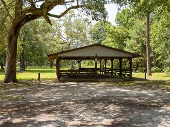 Wooden open air structure with wooden picnic tables underneath. Gravel drive in the foreground. Green trees in the background.