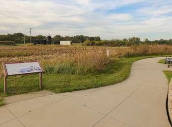 Concrete pathway meandering through grass and shrub meadow with interpretive waysides and metal benches