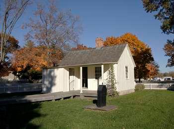 A cottage's roofed back porch has a lean-to with a vine-covered trellis, and overlooks a black antique water pump in the back yard.