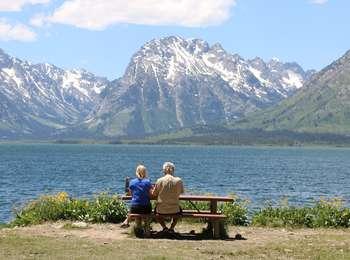 Two people sitting at a picnic table at the Colter Bay picnic area looking across Jackson Lake toward Bivouac Peak.