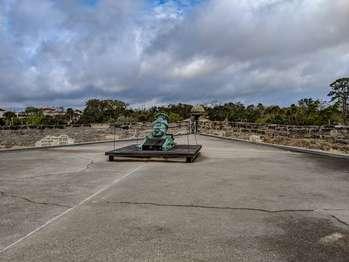 When viewed from above, as from a bird’s eye, the Castillo is square in shape, with four diamond-shaped battlements extending from each corner. These diamond shaped bastions are connected by the gundeck. The San Pablo Bastion is located on the northwest corner of the Castillo. The view from this bastion includes the moat, the north green of the Castillo, and the City’s parking garage. The historic city can also be seen from this location. The bastion has low walls, about 5 feet tall. Each side of the diamond-shaped structure is unique. Facing the bastion from the gundeck, going from left to right: At the left corner is a two step platform that wraps around the corner. The walls have cutouts about three feet wide by three feet high, stopping at two feet height above the floor. There is a cutout in the south facing wall with a wayside exhibit in front of it and another cutout to the right of it. At the northwest corner is a sentry box—a cylindrical shaped structure, with a domed top. It is closed to the public by a wooden barricade. The west facing wall has one cutout. In the center of the bastion is a 9 foot square, low wooden platform. A rope stretches between metal posts at each corner. In the center of the platform is another wooden platform on which sits a 15-inch diameter bronze mortar. The bronze has weathered into a pale green patina color. The tube-shaped barrel of the mortar is tilted up into the air at about a 45-degree angle. Its base is connected, crossways, to a thick bronze rod. This rod is secured to the wooden platform by thick metal strips with rounded metal knobs. The front of the mortar platform has a raised wooded lip with metal strips securing it on each end. The mortar barrel has ornate bronze sculptures. At the center of the barrel is a coat of arms. At the top, are two curved handles in a dolphin shape.