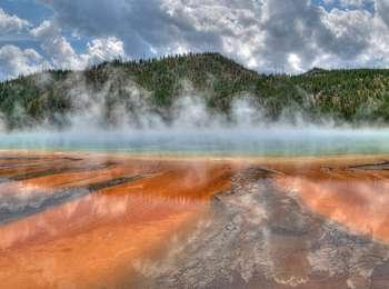 Orange streamers extend from a steaming blue pool in front of a forested hill