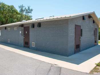 A grey stone building on cement with a brown roof and brown doors. A brown restroom sign hangs by a door. Green trees and blue sky in the background.