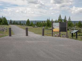 Signs and wooden pillars mark the start of a wide trail across an open meadow.