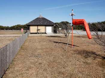 An orange windsock blows in the wind to the right of the wooden fence denoting the entrance to the Ocracoke Airstrip and the pilot facility in the background.