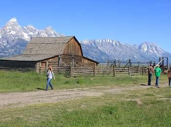 Visitors in front of John and Bertha Moulton barn with a corral to the right. The Teton Range including the Grand Teton to the left are in the distance. 