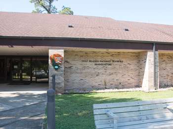 A light brown brick building with a dark brown roof. The National Park Service orange and green arrowhead logo on the building. Black text reads,