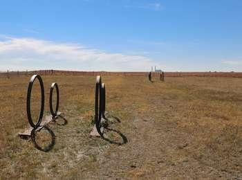 Metal wagon wheel sculptures mark faint Oregon Trail swales in a dry, grass meadow that stretches to the horizon