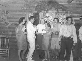 Several young men and women enjoy a costume party inside a log cabin, with balloons and other decorations on the walls.