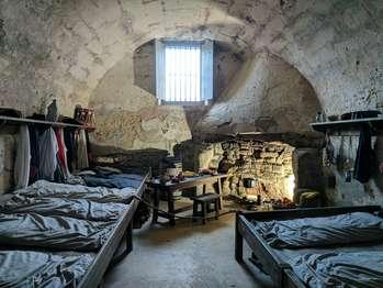 Vaulted ceiling room with bunks connected to the side walls with 8 mattresses. shelves holding a drum, lantern, shoes, tricorn hats, bowls, and pottery. a table with food on it is in the center of the room and behind the table is a fireplace with cooking implements in it