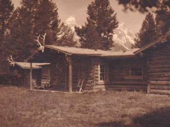A large log cabin decorated with elk antlers on the eaves stands among tall pine trees, with snow-capped peaks in the distance.