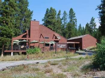 Approach to Leeks Pizzeria. Paved path leading to two red-brown buildings separated by a breezeway. A patio with tables wraps around the main building.