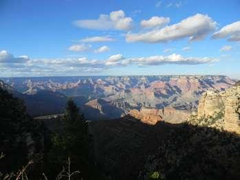 A late afternoon sun casts the canyon in warm tones where ridges are highlighted and valleys hidden in shadow. High overhead white and grey clouds dot the sky.