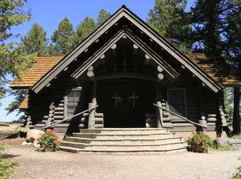 Front entrance to the Chapel of the Sacred Heart. Brown log cabin with shake roof, stairs leading up to double door entrance with crosses on them.