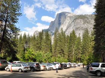 Rows of cars fill a parking lot surrounded by trees. A large granite rock feature rises up behind treeline. 