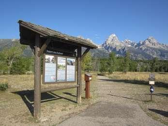 Looking past the Taggart Lake Trailhead along a dirt trail through a sagebrush meadow and the Grand Teton in the distance.