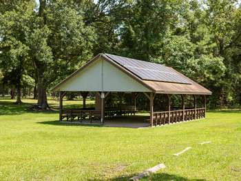 Wooden open air structure with solar panels on the roof and picnic tables underneath. Green grass in the foreground. Green trees in the background.