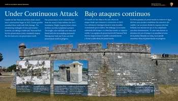 Along the top half of the slanted panel, white text against a blue background in English and Spanish reads:  Under Continuous Attack. Castillo de San Marcos has been under attack since construction began in 1672. Enemy gunfire assaulted these walls with little damage. The unrelenting onslaught of time and weather, however, are taking a visible toll. National Park Service preservation crews constantly inspect the fort and carry out preservation work.  Past preservation work removed water from the moat to help stabilize the fortâ€™s foundation. Bright coquina shows where workers recently completed a project. The bright color will fade over time and blend with the surrounding stonework. During your visit, see if you can find preservation work in progress.  End of Text  On the lower half of the panel, a large color photo of the fortâ€™s grey, wedge-shaped, bastion. In front is a full view of the wall with 3 cut-outs for cannon positions on top.  On the left is a small round tower. Set behind this wall, a partial view of the fort curtain wall with a single rectangular window. Above, a black cannon pokes out of a gun port.  Below, are 2 inset square color photos.  At the left, a close-up view of a section of mottled grey coquina stone that is revealed as the historic layer of white plaster weathers away.  To the right, a close-up view of two, 4-sided tall chimneys on the fortâ€™s gun deck. Near the top of each chimney, on each of its 4 sides, are rectangular-shaped openings for smoke to vent out. The chimney at the left is a dark grey and white. The upper half of the chimney at the right is beige new coquina blocks with white mortar; its lower portion is light grey.  Below, against a translucent blue background, white text in English and Spanish reads:  Preservation efforts stabilize this coquina chimney, but the outer plaster layer has not been replaced.  End of Text  At the lower right corner of the photo, a snaking crack lines the fortâ€™s grey faÃ§ade and coquina is wearing away below a scupper. White text against a translucent blue background in English and Spanish reads:  The wall in front of you shows more than a century of water damage (as seen in 2016).  Beyond the panel are views of the fortâ€™s outer wall with long, jagged vertical cracks in the light grey and white faÃ§ade. Directly below the top of the wall are small scuppers that drain water from the gun deck. The wall below the scuppers is eroded and discolored to a shade of brown.  Directly in front of the panel and down 4 feet is the grass and dirt covered way. Set on a concrete pad is a 3-foot high pedestal sign that reads â€œKeep off the moat stairs.â€,   [End of Message]
