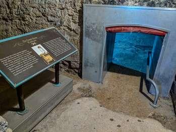 The Powder Magazine rooms are accessed through the Artillery Complex. The entrance to the Power Magazine area is on the northeast wall of the interior room of the Artillery Complex, to the right of the Ignacio Daza portrait. This entry way is 6 feet 5 inches tall and three feet wide. The first room is long and narrow stretching off to your left. The entrance to the Powder Magazine Room is straight ahead. In this first room, to the left, an exhibit blocks off part of the room. On the exhibit, in English and Spanish, reads, The Powder Room Exhibit. An illustration shows a section cut through the magazine room. It shows a narrow opening up through the roof, to the gun deck level of the Castillo. The text reads, “You can enter the former powder magazine through this doorway. Built in 1675, it is one of the oldest rooms in the fort. It was originally built to store gunpowder but soon proved too damp. During the 1702 siege, the room was used as a trash pit and then sealed closed. When reopened over 100 years later, animal bones found inside inspired many local myths, one claiming the bones belonged to lovers buried alive inside the Castillo.â€, On the wall, to right of the exhibit, you can enter the next room by crawling through a metal, 3 foot deep, entry tunnel. The tunnel is 3 feet high and 2 feet wide. Once you have knelt down, there is a metal handrail that curves up from the ground at 12 inches on your right. The handrail continues through the tunnel. The top of the tunnel is padded, but please watch your head. Once you have passed through the tunnel, you can stand up. This is the Powder Magazine room. It is 20 feet by 20 feet with an arched ceiling, about 7 feet high. The room is empty. The floor is uneven with loose crunchy pebbles. To exit the room, you must return through the metal tunnel as there are no other exits.