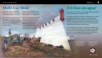At the left and right sides of the panel, against a color illustration of light blue sky with pale pink clouds, black text in English and Spanish reads:.  Multi-Use Moat. It smells and sounds like a farmyard. Mooing cows, clucking chickens, and snorting pigs are penned in the moat.  The 1702 British siege made staying in town dangerous. Townspeople moved into the fort for protection, bringing their livestock with them. Farmers brought their recent harvest to extend the storeroom rations. For more than a month, nearly 1500 people crowded into the fort, relying on these provisions until the siege ended.  This wide, dry ditch made the Castillo less vulnerable to infantry attacks. Combined with other defenses, the moat protected the fortâ€™s lower walls from cannon fire. The uncovered expanse also exposed would-be attackers to soldiers firing from the fortâ€™s gun deck.   End of Text  Between the English and Spanish text, a wall of the white, wedge-shaped fort bastion recedes back into the distance. The ends of several cannons poke out of square-shaped cut outs called embrasures along the upper portion of the fortâ€™s wall.  Below the fort is a muddy area where 5 cows, 3 with horns; 6 brown and black pigs; and 13 chickens meander.  A man in a blue shirt, white pants, and cap sticks a pitchfork into the back of a straw-filled blue cart. Chickens peck on the straw that is already on the ground.  On the left, on top of the coquina stone wall a man in a knee-length brown coat, knee-high brown boots, and black hat leans against a long brown musket.   To the right, white text caption in English and Spanish reads:  The moat was filled with water from 1938 until 1996, when the National Park Service drained it to reflect its original appearance.  At the bottom right, a small circle divided into 4, triangular wedges. Each wedge has a portion of a flag and date range for an important period in the fort's history. 1 wedge is bright, indicating the time period of the panel story and the other three wedges are muted. From the top, moving clockwise: Great Britain: 1763-1784; Spain: 1784 to 1821; the United States: 1821-1900; and Spain: 1672-1763, is bright.  Below the circle, white text in English and Spanish reads: First Spanish Military Era.  Beyond the panel, a grassy area in between the 6-foot high stone wall to the left, and the fortâ€™s 30-foot high grey stone wall to the right is the moat. Ahead, a brown wooden walkway set on 6 Â½ to 4-foot high stone pylons that run from the wall at the left to the fortâ€™s entranceway at the right.  [End of message]