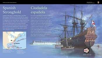 On the left, against a deep blue background, light blue and white text in English and Spanish reads:  Spanish Stronghold. Castillo de San Marcos symbolizes Spainâ€™s vast New World empire. Built between 1672 and 1695, the coquina fort replaced a series of wooden forts that had protected St. Augustine for more than 100 years. Improvements in the mid-1700s helped the Castillo, with its soldiers and guns, keep pirates and the British at bay. Spain controlled Florida, and the Castillo helped keep their enemies away from the Gulf Stream shipping lanes. Spanish treasure fleets used these routes to carry sugar, tobacco, pearls, silver, and gold to Europe.  End of Text  Below on the left, a small, rectangular map: a British flag marks the beige upper right, the eastern seaboard of the United States above the state of Florida. A French flag marks the off-white center part of North America. Below, a Spanish flag marks the light brown area that includes the rest of North America, Central America, Caribbean islands, and the northern coast of South America. To the right, against a light blue background, curving, red lines with black arrows above and below, mark shipping routes to and from Spain. Black dots mark city ports: La Habana, Santo Domingo, San Juan, Cartagena, Panama, Portobello, Veracruz, and Acapulco.  Below, white text in English and Spanish reads:  Shipping routes, the major roadways of their day, connected continents and cultures.  End of Text  On the right side of the panel, against a blue sky is a dark brown wooden ship anchored facing the Castillo de San Marcos fort. Dozens of brown ropes run from the top of its 3 tall masts to the deck. Attached to a brown flagpole at the shipâ€™s high carved stern, or rear is a white first Spanish Military Era flag with 2 red crossing lines in the center. In the water, to the left of the ship are 2 small white boats with single masts and men in white shirts and pants with red sashes at the waist.   At the bottom right, a small circle divided into 4, triangular wedges. Each wedge has a portion of a flag and date range for an important period in the fort's history. 1 wedge is bright, indicating the time period of the panel story and the other three wedges are muted. From the top, moving clockwise: Great Britain: 1763-1784; Spain: 1784 to 1821; the United States: 1821-1900; and Spain: 1672-1763, is bright.  Below the circle, black text in English and Spanish reads: First Spanish Military Era.  Directly beyond the panel, a low stone wall; to the left, a light grey field-stone walkway with low stone walls on either side. Ahead, and beyond the low walls, views of the Matanzas River.  For additional information, turn to your left and move ahead to a park ranger station in a one-story brown wooden building.  [End of message]