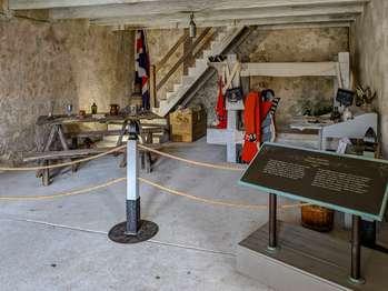 The British Room is accessed from the courtyard or the Second Spanish Period room. This room shows how British soldiers remodeled the Castillo’s rooms during their occupation. This room is described based on entering from the courtyard. A wooden door opens into the room. Above the door is a barred window with a wooden shutter that opens inward. Barred windows are on either side of the door. The room is about 20 feet wide and 30 feet long. A flat, wooden beamed loft is at a 10 foot height, the 17-foot arched ceiling is visible above the wooden second floor as you stand just inside the doorway. Wooden stairs along the back wall lead up to a loft. On the right is an entrance to the Second Spanish Room. Standing at the entrance to the room, moving from left to right, the exhibits, in English and Spanish, include: Inside a Casemate Exhibit. A bronze tactile model shows a cross section of how this casemate would have looked during the British Period. Sir Frederick Haldimand. 1718 – 1791. Text explains that a Swiss mercenary hired to fight for England became a colonel, and eventually, a knight. Haldimand was posted here from 1769 to 1770. In an illustration, he wears a black triangular hat with jaw length gray hair. He has thick black eyebrows, brown eyes, and a broad nose. He is wearing a red jacket with blue trim with a silver epaulet on the right shoulder. He wears a white vest with silver buttons and tan breeches. His white ruffled shirt has a black collar. Around his neck, he wears a flat, silver medallion, with an engraving of a coat of arms. Two inches of ruffled white lace shows at his wrists. A red sash is tied at his waist. He wears knee high black boots. He holds a silver sword with a gold tassel on the hilt. The living area is roped off. On the left, is a table with a checkers game and goblets. At the far right corner is a white wood bunk bed, with uniforms hanging on pegs. Nearby, there is a gray desk with a flip-down top, and a gray chair. Next to it, on a gray wooden stand, is a metal tub, with a rag hanging over the edge. Close Quarters Exhibit. The text explains that multiple bunk beds occupied each room. Four soldiers would have slept on a bunk bed. Two on each bunk of the bed. Great Britain Takes Control Exhibit. A photo shows re-enactors in this room, wearing red coat uniforms, with black triangular hats. An illustration shows a man in the same attire. A historic map of St. Augustine. A portrait of an older man, in a uniform with many badges and epaulets. The text explains that Great Britain takes over the Castillo in 1763. Within a few years, the Revolutionary War led the British to strengthen the fort’s defenses.