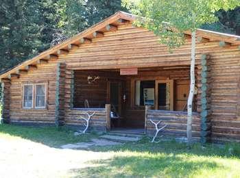 Murie Residence cabin tan logs with an covered inset porch. Ends of logs painted green. Aspen tree in front and elk antlers.