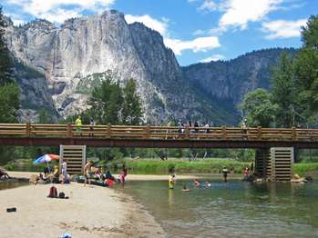 People walk over the swinging bridge while people play in the water below it. A tall, very thin waterfall is in the distance.