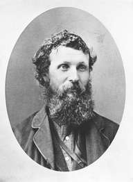 A black and white portrait photo shows a man with tousled short hair, an unkempt beard and mustache, wearing a button-up shirt and rugged coat.