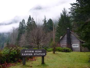 A cabin with a stone chimney among trees and mist. Sign in front reads
